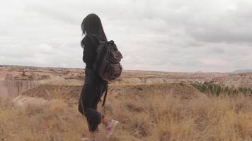 Side tracking view pretty tourist woman with hiking retro backpack walking alone through valley wild nature enjoying a hiking adventure alone in cappadocia in cloudy day, Turkey travel and explore video