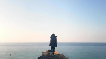 Female person walks closer to edge of the cliff stops and looks around with cast sea background video