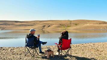 Young caucasian Couple sit on foldable chairs by lake drink coffee outdoors in scenic sunny location while travel with tranquil lake panoramic background . Romantic couple outdoors concept
