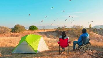 Young caucasian romantic couple sit and enjoy balloons in sky on sunny morning at sunrise while camping .Travel cappadocia relaxing holidays destination