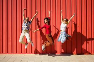 Playful young diverse friends jumping on street against red wall photo