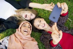 Cheerful diverse girlfriends laughing and taking selfie lying on lawn in park photo