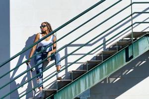 joven mujer negra con trenzas afro y gafas de sol en una escalera urbana, llevando una patineta. foto