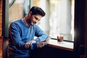 Smiling man with blue sweater looking at his smartphone in a modern pub.