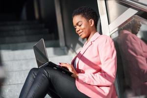 mujer de negocios negra sonriente sentada en escalones urbanos trabajando con una computadora portátil foto