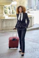 Black woman smiling and carrying a rolling suitcase in urban background photo