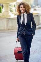 mujer de negocios negra con cabello afro caminando con una maleta rodante. foto