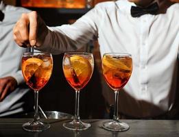 three glass glasses with an aperol spritz cocktail are on bar. bartender in white shirt, bow tie is stirring one of aperitifs. concept - people, drinks, entertainment, recreation. free space for text.