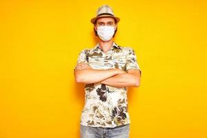 male tourist traveler in medical mask stands with his arms crossed on his chest with serious look. isolated on yellow studio background. concept - vacation, vaccination, protection from coronavirus photo