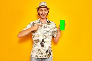 smiling male tourist traveler points with index finger at green blank phone screen to insert an advertisement. isolated on studio yellow background. concept - people, technology, advertising, vacation photo