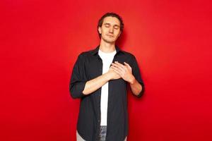 man with eyes closed holds both hands on chest showing heart pain, heart problems, needs medical help. dressed in casual clothes, isolated red studio background. concept - people, medicine, gratitude photo