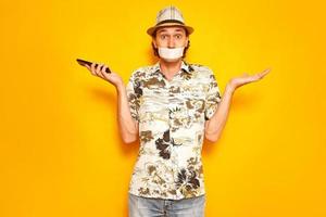 male tourist traveler with duct tape over mouth cannot talk on phone, does not understand problems with communication abroad. Isolated on studio yellow background. concept - people, travel, technology photo