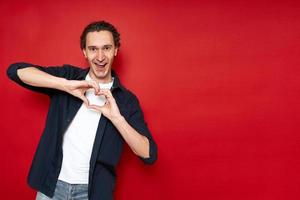 cheerful excited man shows heart sign with hands in front of his chest, confesses his love with his mouth open. isolated on red background copy space. concept - romance, relationships, valentine's day photo