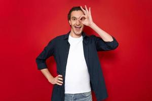 excited man with mouth open holds his hand in form of an ok sign near eye and looks through his fingers. dressed in casual clothes isolated on red studio background. concept - people, approval, sale photo