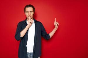 serious man holds his index finger to his lips with his other hand, points to an empty area for advertising text. secret sale taboo. isolated on red background. concept - people, mystery, black Friday photo