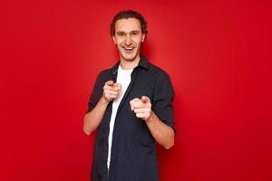 smiling, excited man points his index fingers forward in front of him. he is dressed in casual clothes, blue shirt, white T-shirt. isolated on red studio background. concept - people, victory, best photo