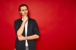 man holds chin his hand in casual clothes blue shirt in thoughtful pose makes difficult choice isolated red background space for advertising text concept - brainstorming, idea, comparison, analysis photo