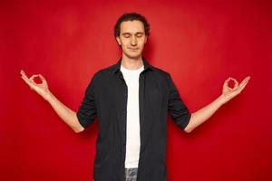 Smiling young brunette man wearing basic casual blue shirt hold hands in yoga gesture, relaxing meditating, trying to calm down isolated on bright red colour background, studio portrait, copy space photo