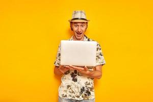 surprised joyful male tourist with laptop in his hands received an email about the win with an enthusiastic expression on his face from seeing favorable offer for discounts. isolated yellow background photo