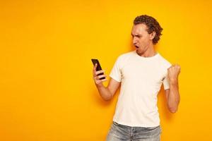 excited man checks his mobile phone celebrating good news with victorious gesture isolated on yellow background in casual clothes. space for text. Concept - communication, people, social networks photo
