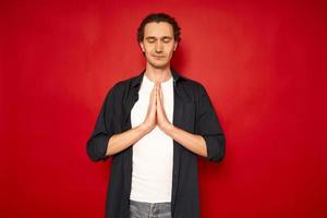 concepto de meditación. un hermoso joven se para en una pose meditativa, disfruta de un ambiente tranquilo, sostiene sus manos en un gesto de oración, aislado contra el espacio de copia de fondo rojo, tiene un sentido de paz interior foto