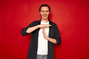 una foto de estudio de un hombre alegre y sonriente que usa ropa informal muestra que el gesto de tiempo de espera necesita parar pide tiempo para descansar después de que el trabajo duro demuestra el signo de la mano de descanso. aislado del concepto de personas de lenguaje corporal de fondo rojo