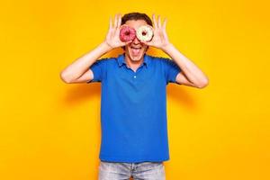 funny young hipster man in casual clothes holds sweet multicolored donuts on his eyes like glasses, binoculars. blue polo isolated on bright yellow background. concept food, holiday, diet, people. photo