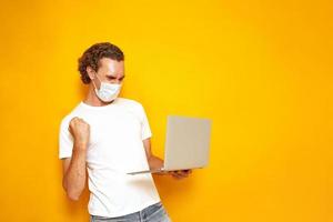 man in casual clothes with laptop in his hands and protective medical mask happy win showing winning gesture. isolated on yellow background with space for text. concept people, victories, advertising photo