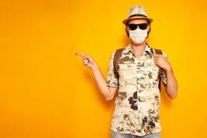 a male tourist in a medical mask isolated on a yellow background points to an empty area with his finger. he is wearing a Hawaiian shirt, a hat, sunglasses, a backpack on his shoulders. space for text photo