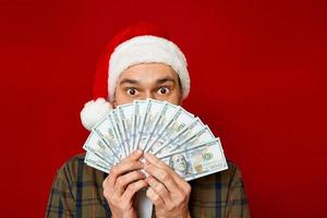 large studio portrait of young man in Christmas hat with wad of money in his hands holding fan of dollars in front of his face in shock from successful lottery win. concept winning, cash back, wealth photo