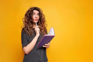 young woman writer in casual clothes thinking about text search inspiration and new ideas putting pencil to her mouth holding notebook in hands notebook for writing stands yellow isolated background photo