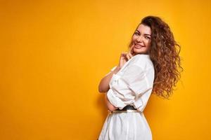 young woman with curly hair, positive facial expression, good mood and joyful smile in casual clothes white shirt stands sideways isolated yellow background holding hand her chin thinking about buying photo