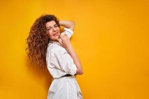 young curly haired woman in white shirt holds her hand to chin and adjusts her hair with thoughtful face stands sideways isolated on yellow background with place for text thinking about right decision photo
