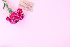 fotografía conceptual de la idea del día de las madres: hermosos claveles florecientes atados por un lazo con una tarjeta de texto kraft aislada en una mesa moderna y brillante, espacio de copia, capa plana, vista superior foto
