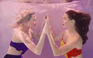 art portrait of two beautiful pretty women holding each other hands underwater on pink background photo