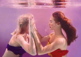 art portrait of two beautiful pretty women holding each other hands underwater on pink background photo