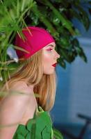 retrato de atlética hermosa mujer rubia atractiva con maquillaje en gorro de baño y traje en la piscina con flores y plantas verdes foto