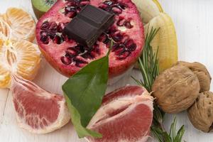 A pomegranate with chocolate, grapefruit, tangerines, walnuts, and banana on a white wooden table photo