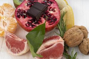 A pomegranate with chocolate, grapefruit, tangerines, walnuts, and banana on a white wooden table photo