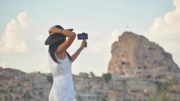 visão estática jovem turista feminina de vestido branco com smartphone em tripé handheld panning e capturar férias em localização cênica