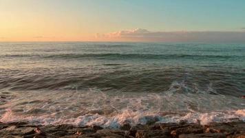 visão estática ondas em tempo real quebram sobre rochas na praia com horizonte e fundo de céu claro em branco azul laranja pôr do sol. panorama tranquilo e romântico da praia video