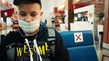 jovem de máscara azul com mochila toma assento vazio no saguão do terminal do aeroporto para aguardar o voo após as restrições de quarentena video