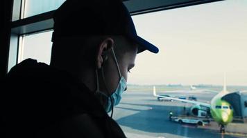 el joven se para cerca de la ventana y espera el vuelo. el tipo escucha música observa a los trabajadores que preparan el avión para el vuelo en la plataforma video