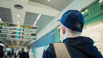 junger mann mit medizinischer maske in kappe steht im flughafenterminal unter menschen, die die informationstafel für das fluggate betrachten video