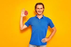 smiling man holds donut in his hand in form of an OK sign. he is dressed in casual clothes, blue polo, jeans. isolated on yellow studio background with space for text. concept - people, food, sweets photo