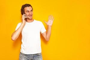 smiling man is talking on phone gesticulating with his raised hand, enjoying communication, dressed in casual clothes. isolated on studio yellow background. concept - people, technology, communication photo