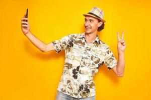 smiling male tourist traveler takes selfie on phone camera showing a victory peace sign with his hand. isolated on a yellow background with space for text. concept - people, communication, technology photo