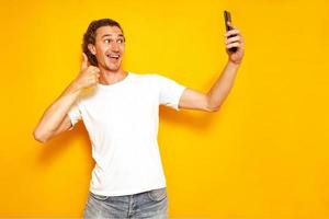 excited man takes selfie on his phone, gives thumbs up sign communicates well via video smartphone smiles isolated on yellow background with space for text. concept - people, technology, communication photo