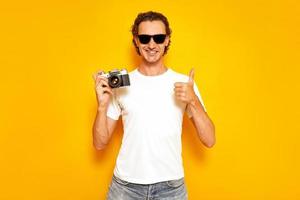 male photographer with retro camera in his hand gives a thumbs up dressed in casual clothes, white T-shirt, jeans, sunglasses isolated on a yellow studio wall. concept - people, creativity, technology photo