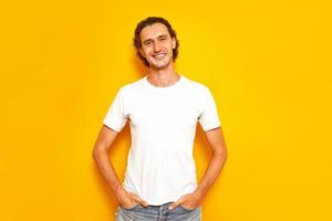 young handsome smiling man in casual white T-shirt standing on an isolated yellow background, keeps his hands in his pockets. an approving expression on face looking into the camera indicates success photo